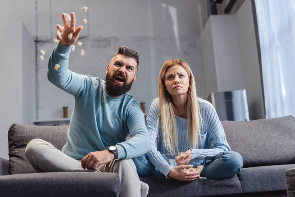 Emotional husband throwing popcorn and sitting on sofa with wife — Stock Photo