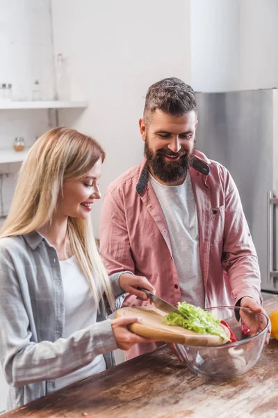 Casal de pé na cozinha e adicionando folhas de salada na tigela — Fotografia de Stock