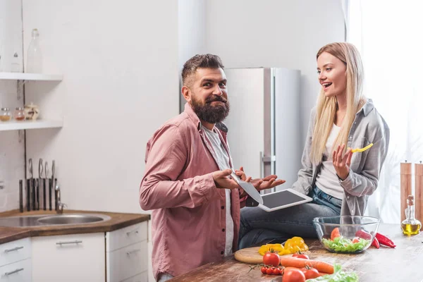 Marido gesticulando e olhando confuso enquanto esposa sentada na mesa com tablet digital — Fotografia de Stock