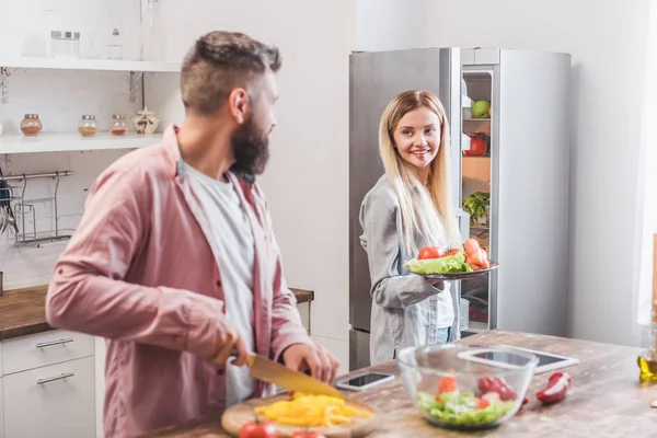 Esposo cortando verduras y esposa de pie cerca del refrigerador - foto de stock