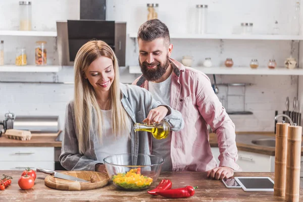Bela esposa de pé com o marido na cozinha e adicionando óleo na salada — Fotografia de Stock