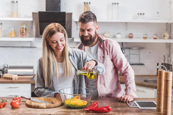 Belle femme debout avec mari dans la cuisine et ajouter de l'huile dans la salade — Photo de stock