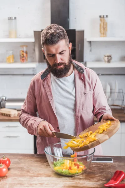 Hombre barbudo guapo agregar pimiento picado a la ensalada en la mesa de la cocina - foto de stock