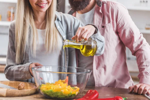 Обрезанный вид улыбающейся женщины, стоящей рядом с мужем и добавляющей масло в салат в миске — стоковое фото