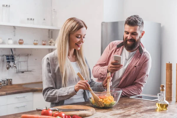 Sonriente barbudo hombre tomando foto de la mujer cocina ensalada en la cocina - foto de stock