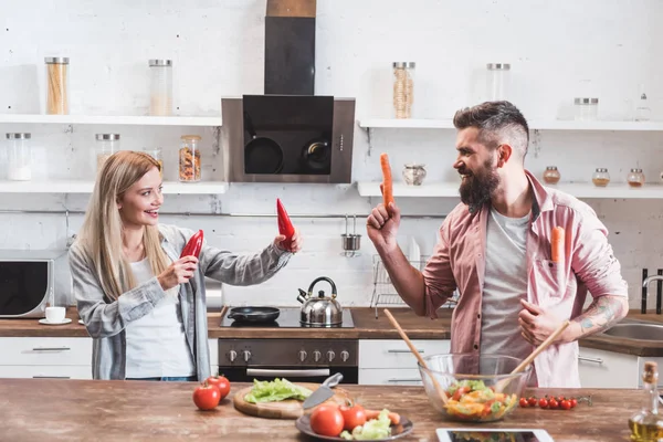 Divertida pareja jugando con verduras mientras cocina la cena en la mesa de madera - foto de stock