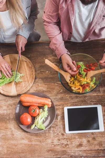 Vista recortada de pareja cocinar ensalada de verduras con tableta digital en la mesa de madera - foto de stock