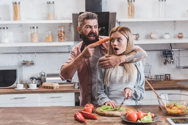 Gracioso hombre poniendo zanahoria cerca de mujer cabeza mientras asustado chica cocinar cena - foto de stock