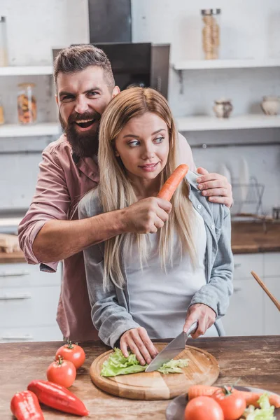 Engraçado homem colocando cenoura perto mulher garganta enquanto menina cozinhar jantar — Fotografia de Stock