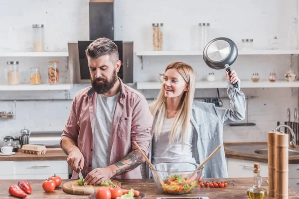 Lustige Mädchen scherzen und heben Metallpfanne für das Schlagen Mann in der Küche — Stockfoto