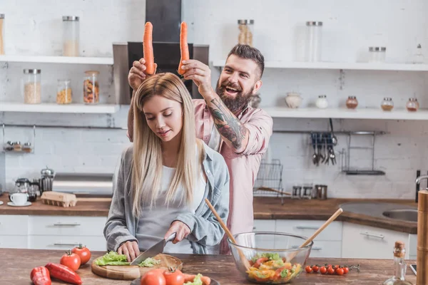 Смешной мужчина кладет морковку на голову женщине, пока девушка готовит ужин — стоковое фото
