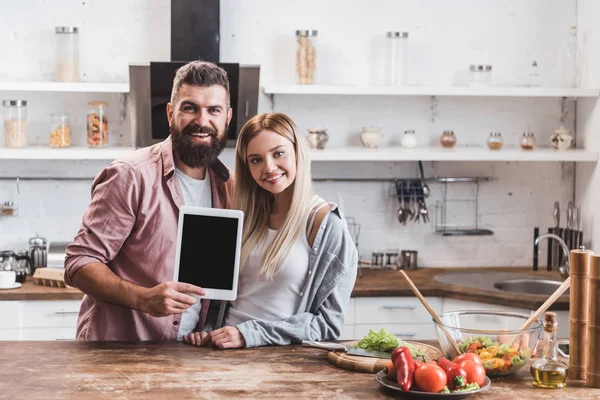 Feliz pareja sosteniendo tableta digital con pantalla en blanco en la mesa de la cocina de madera - foto de stock
