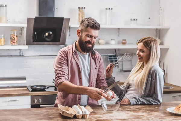 Bell'uomo barbuto rompere le uova al tavolo di legno, mentre la donna in possesso di frusta palloncino — Foto stock