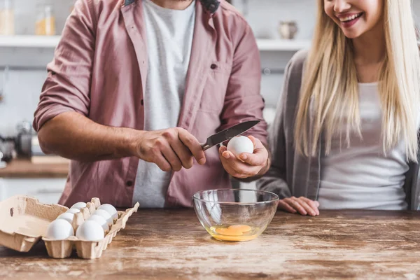 Обрезанный вид улыбающейся женщины и мужчины, разбивающих яйца в миске за кухонным столом — стоковое фото