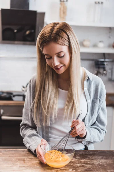 Rubia joven sonriente mujer batiendo huevos en cuenco de cristal en la mesa de madera - foto de stock