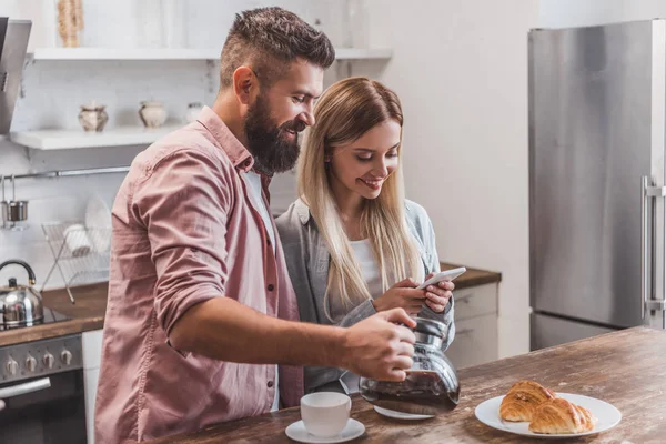 Fröhliches Paar frühstückt und nutzt Smartphone in der Küche — Stockfoto