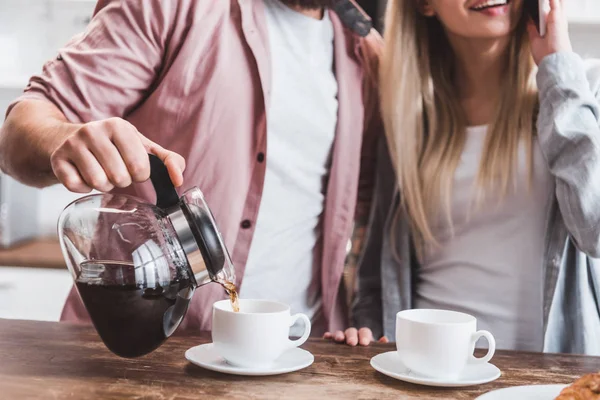 Vista recortada del hombre vertiendo café en la taza mientras que la mujer habla en el teléfono inteligente - foto de stock