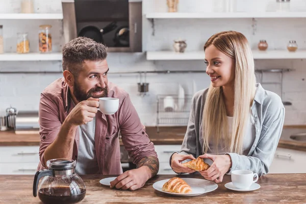 Junges fröhliches Paar isst Croissants und trinkt Kaffee zum Frühstück — Stockfoto