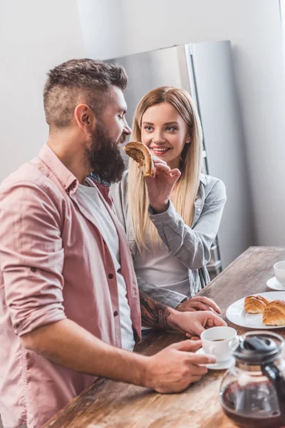 Lächelnde blonde Frau füttert bärtigen Freund morgens mit Croissant — Stockfoto