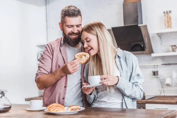 Bearded man feeding girlfriend with croissant at kitchen — Stock Photo