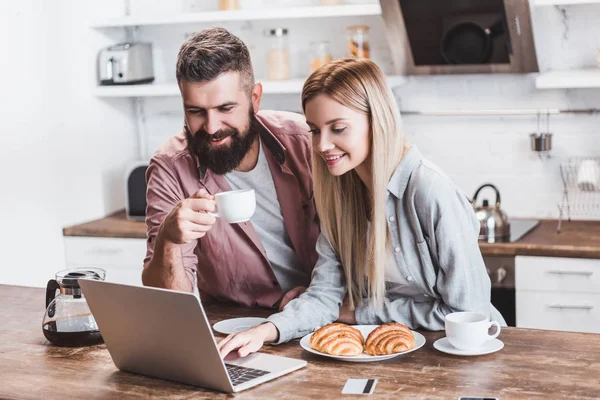 Mujer joven usando el ordenador portátil en la mesa de la cocina, mientras que el hombre sosteniendo taza blanca - foto de stock