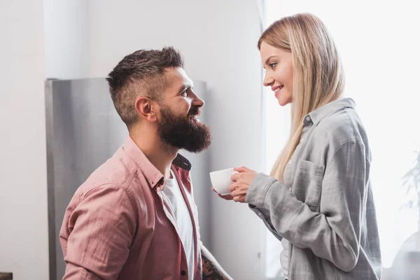 Souriant femme blonde tenant tasse de café tout en regardant petit ami — Photo de stock