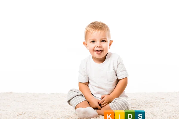 Lachender Kleinkind Junge sitzt auf Teppich neben farbigen Würfeln mit Buchstaben isoliert auf weiß — Stockfoto