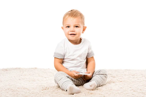 Glücklicher Kleinkind Junge sitzt auf Teppich und hält Smartphone isoliert auf weiß — Stockfoto