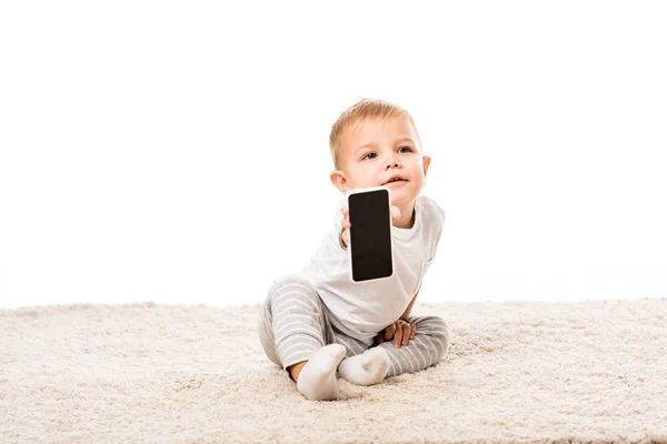 Nice toddler boy sitting on carpet and showing smartphone screen isolated on white — Stock Photo