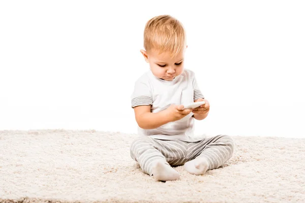 Adorabile bambino con smartphone seduto su tappeto isolato su bianco — Foto stock
