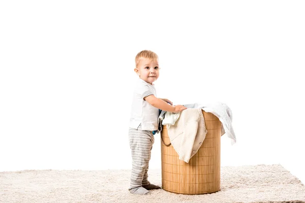 Niño pequeño de pie frente a la cesta de ropa de mimbre y sonriendo aislado en blanco - foto de stock