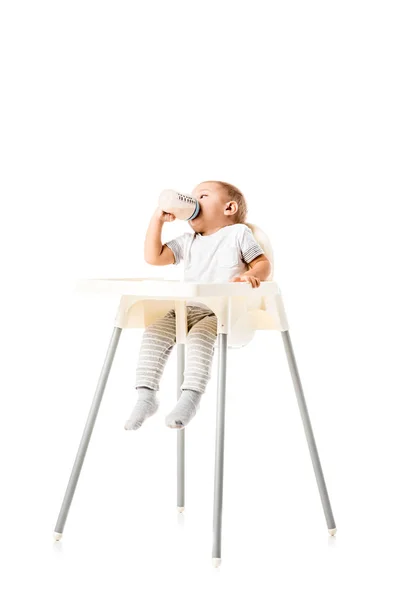 Cute toddler boy drinking from baby bottle and sitting in highchair isolated on white — Stock Photo