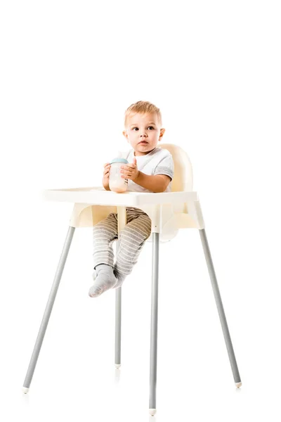 Adorable toddler boy holding baby bottle and sitting in highchair isolated on white — Stock Photo