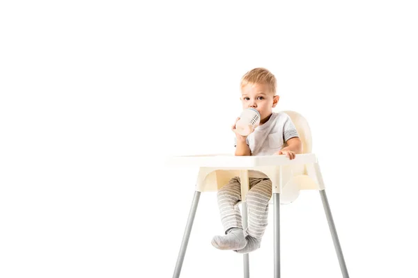 Entzückende Kleinkind Junge sitzt im Hochstuhl und trinkt aus Babyflasche isoliert auf weiß — Stockfoto