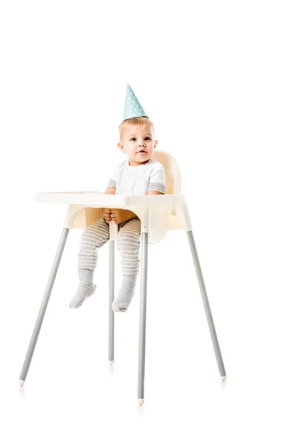 Adorable toddler boy with blue party hat on head sitting in highchair isolated on white — Stock Photo