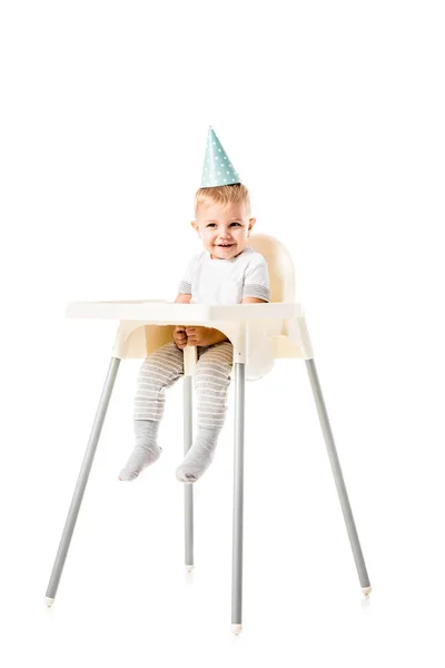 Adorable toddler boy with blue party hat on head smiling and sitting in highchair isolated on white — Stock Photo