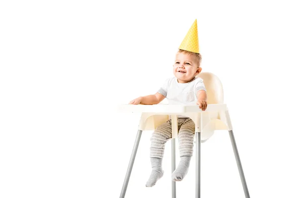 Niño feliz con sombrero de fiesta amarillo en la cabeza sentado en trona y sonriendo aislado en blanco - foto de stock