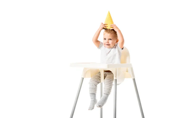Милый малыш сидит в высокий стульчик и положить желтую шляпу партии на голову изолированы на белом — стоковое фото