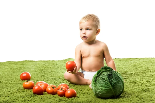Adorável criança menino segurando tomate e sentado no tapete verde com legumes isolados em branco — Fotografia de Stock