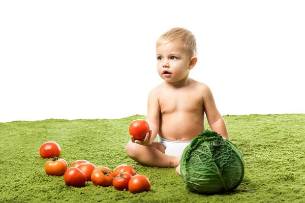 Adorable niño sosteniendo tomate y sentado en alfombra verde con verduras aisladas en blanco — Stock Photo