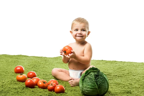Menino sorrindo criança segurando tomate e sentado no tapete verde perto de legumes isolados em branco — Fotografia de Stock