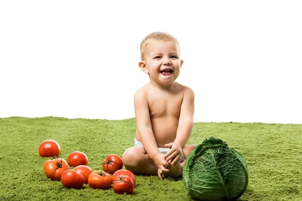 Petit garçon souriant assis avec des tomates et du chou sur le tapis vert isolé sur blanc — Photo de stock