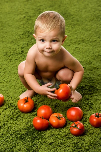 Vista da vicino del bambino sorridente seduto su hunkers e che tiene il pomodoro sul tappeto verde — Foto stock