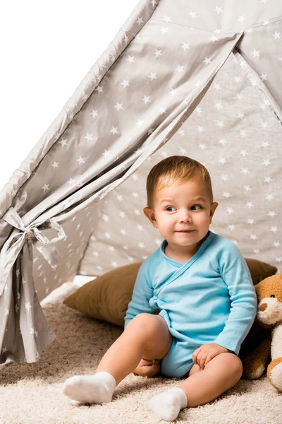 Cute toddler boy smiling and sitting in baby wigwam with pillow and teddy bear isolated on white — Stock Photo
