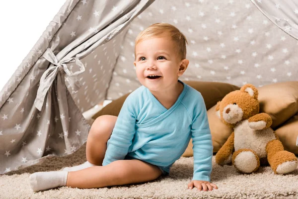 Toddler boy sitting in baby wigwam with pillow and teddy bear and laughing isolated on white — Stock Photo