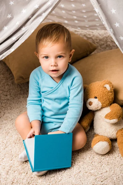 Toddler boy holding blue book, sitting in baby wigwam and looking at camera — Stock Photo