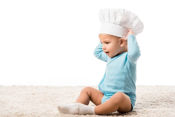 Toddler boy in chefs hat with hands on head sitting on carpet isolated on white — Stock Photo