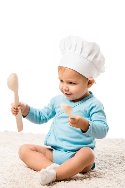 Niño en sombrero de chefs sentado en la alfombra, sosteniendo dos cucharas de madera grandes y sonriendo aislado en blanco - foto de stock