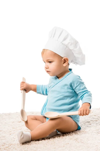 Close up view of toddler boy in bodysuit and chefs hat sitting on carpet and holding two big wooden spoons isolated on white — Stock Photo