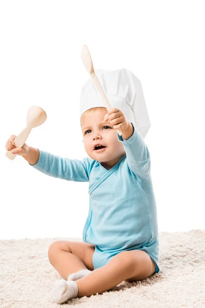 Малыш в шляпе шеф-повара сидит на ковре и держит две большие деревянные ложки изолированы на белом — стоковое фото